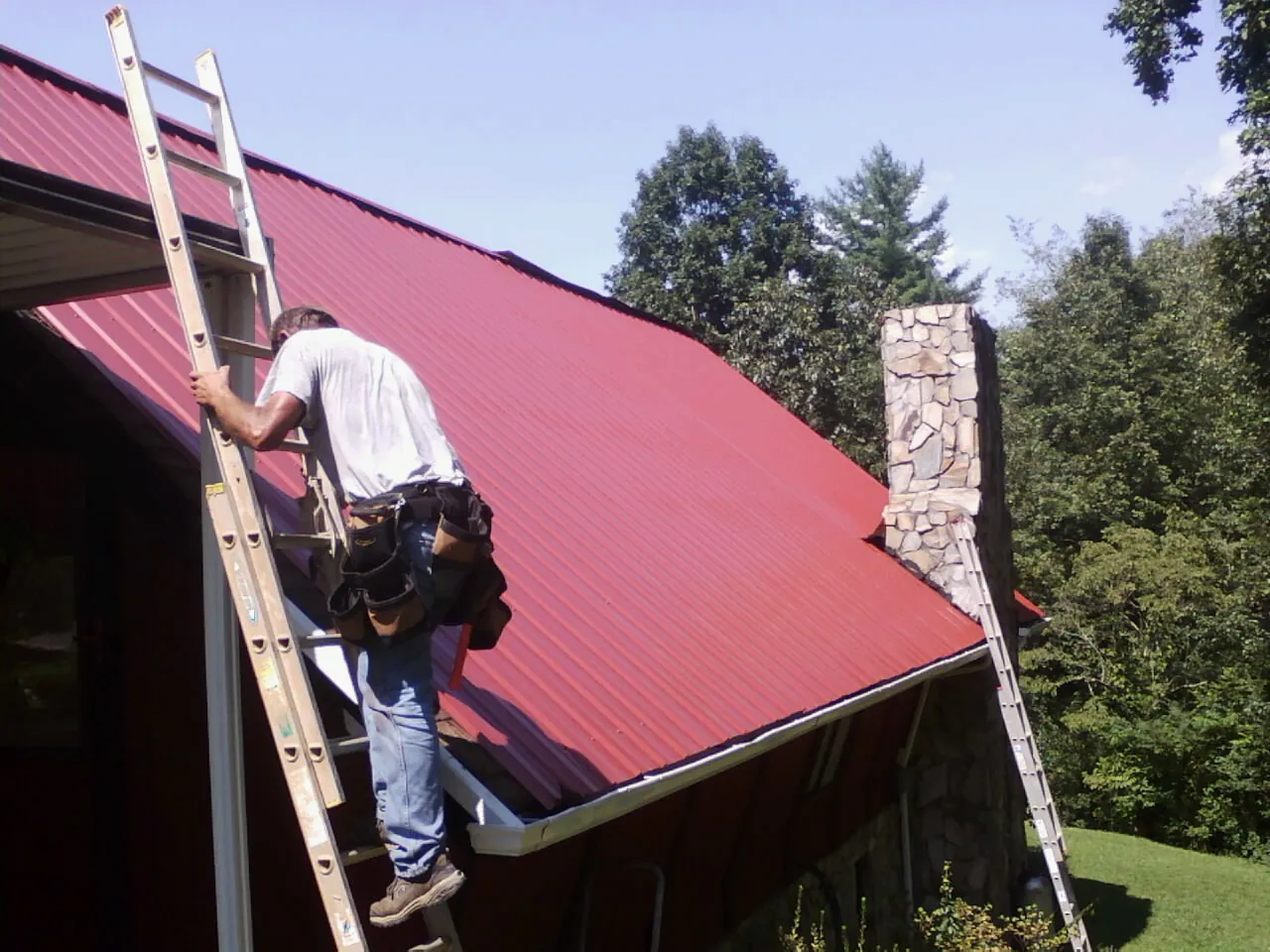 Roofer on a ladder beside a red metal roof and stone chimney — Soffit & Fascia Repair by Roofing Brothers in Lake Elmo